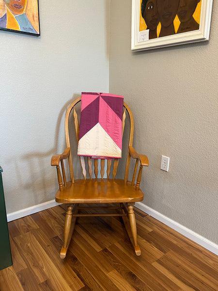 An image of Nemo and Stithc's pink and purple handmade quilt for sale, folded atop a wooden rocking chair in the corner of a room. This quilt is a great handmade baby gift and a wonderful gift for new moms.
