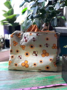 A large pouch stands upright on a green wooden table. The pouch is patterned with Yellow hexagons, bumble bees and sunflowers. The inside fabric peeks up and is hexagons.  There is a natural plant background.
